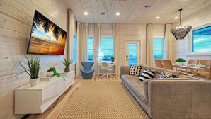 A modern living room with a sofa, dining table, and chairs, a TV mounted on the wall, and large windows with curtains. - Private Pool, Fire Pit, Beach Boardwalk Access, Views The Madison by Avantstay (Port Aransas)