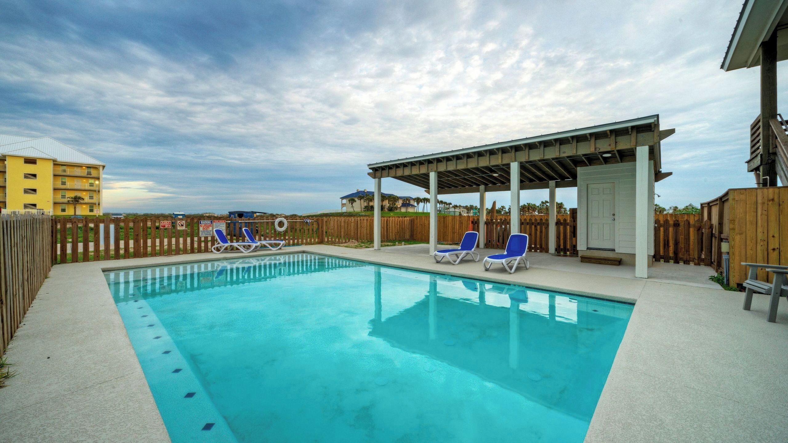 A swimming pool with lounge chairs, a covered seating area, and a wooden fence.