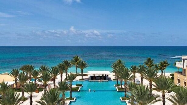 A resort pool with a view of the ocean, surrounded by palm trees and a clear sky.