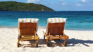 Two wooden beach chairs with striped covers on a sandy beach, facing a calm sea and a green hill in the background.