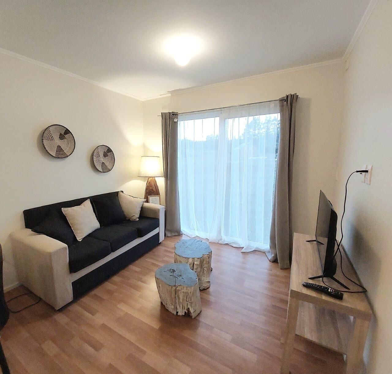 A living room with a black sofa, two round side tables, a TV on a stand, and a large window with curtains.