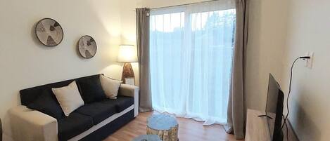 A living room with a black sofa, two round side tables, a TV on a stand, and a large window with curtains.