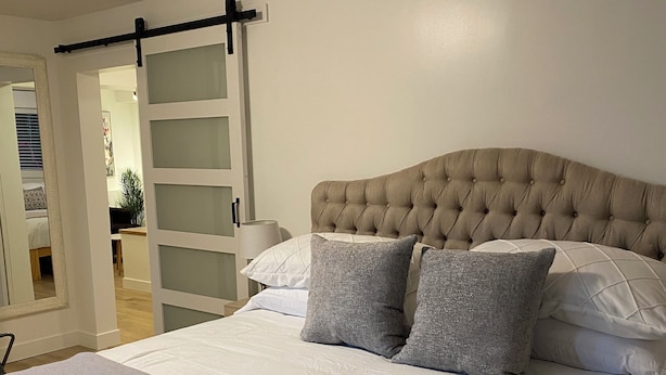 A bedroom with a tufted headboard, a bed with white linens and two grey pillows, a grey throw blanket, and a sliding glass door leading to a bathroom.