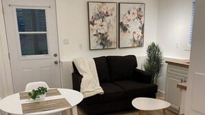 A modern living room with a black sofa, white coffee table, and a dining area with a white table and chairs. There are two framed floral paintings on the wall and a potted plant.