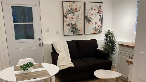 Una sala moderna con un sofá negro, una mesa de centro blanca y un comedor con una mesa y sillas blancas. Hay dos cuadros con motivos florales en la pared y una planta en maceta.