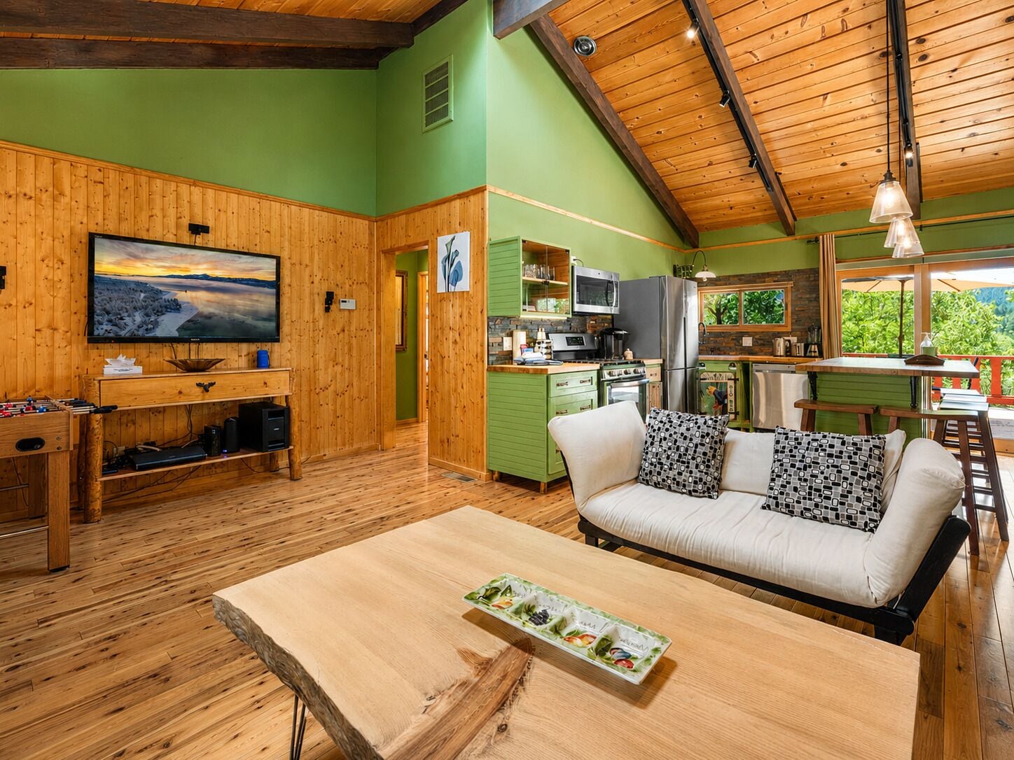A cozy cabin interior with a wooden floor, a sofa with patterned cushions, a dining table, a kitchen area with modern appliances, and a television displaying a scenic view.