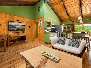 A cozy cabin interior with a wooden floor, a sofa with patterned cushions, a dining table, a kitchen area with modern appliances, and a television displaying a scenic view. - Pinecone Ridge (Big Bear Lake)