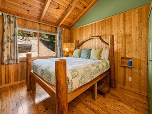A wooden-framed bed with a floral bedspread in a cabin-style room with wooden walls and a window overlooking a landscaped area. - Pinecone Ridge (Big Bear Lake)
