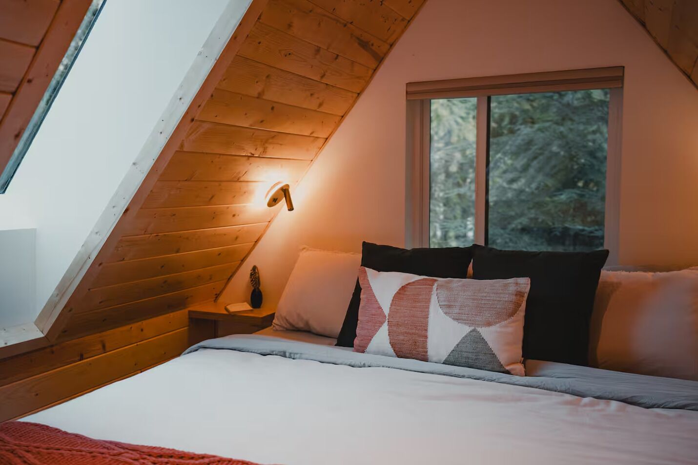 A cozy bedroom with a sloped ceiling, a bed with multiple pillows, a bedside table with a lamp, and a window with a view of trees.