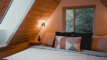 Un dormitorio acogedor con techo inclinado, una cama con varias almohadas, una mesita de noche con lámpara y una ventana con vista a árboles.