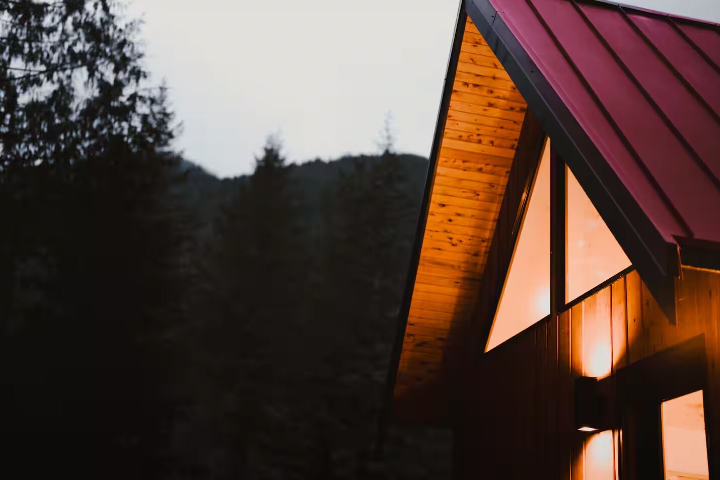 A wooden cabin with a red roof, large windows, and a view of a forested mountain range.