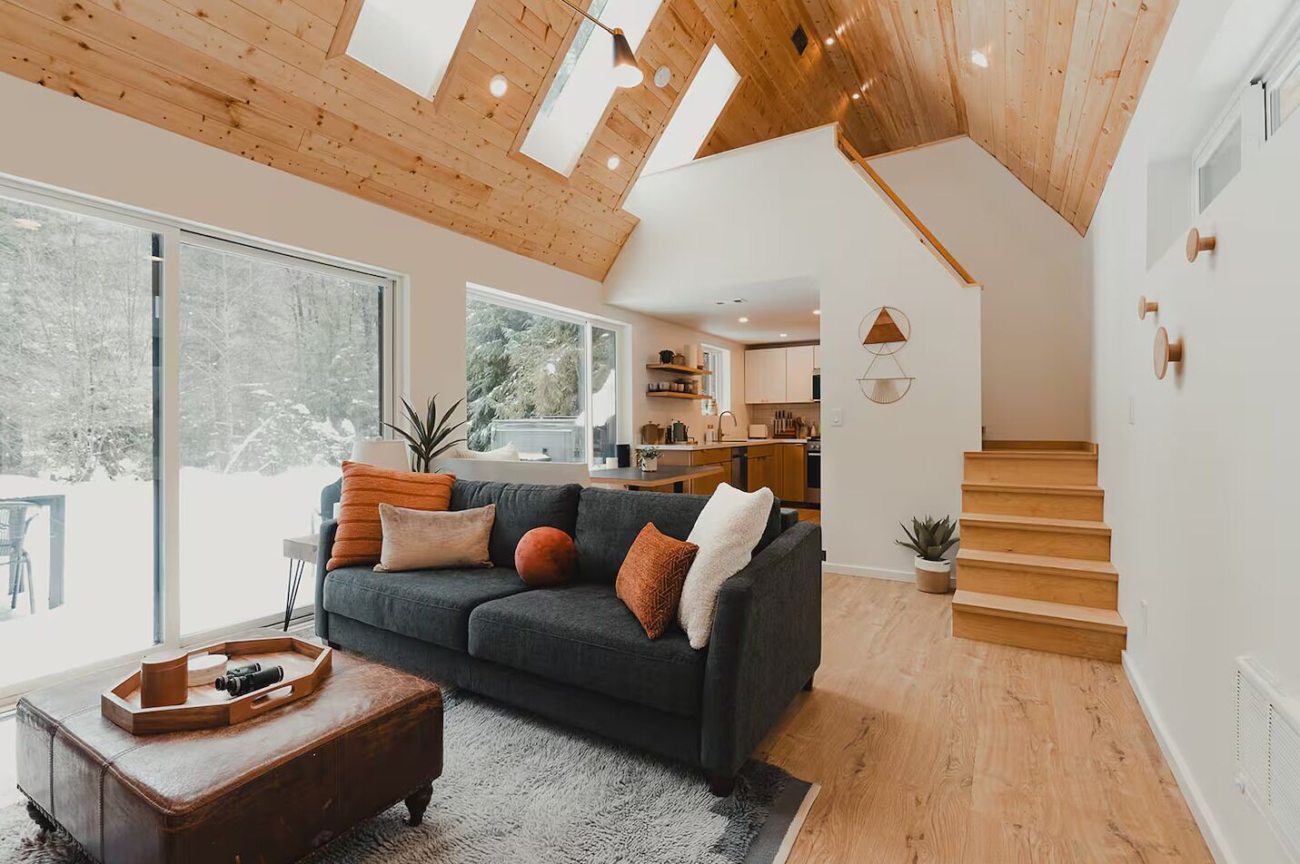 A modern living room with a dark grey sofa, wooden flooring, and a large window offering a view of a snowy landscape.