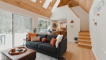 A modern living room with a dark grey sofa, wooden flooring, and a large window offering a view of a snowy landscape.