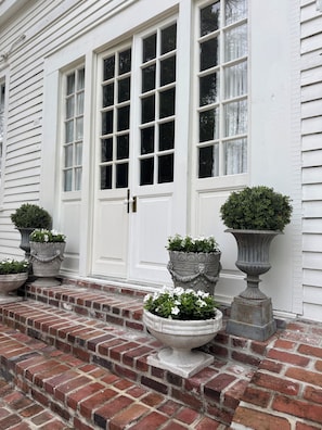 Un bâtiment blanc avec une porte d’entrée, trois grands jardinières ornées de plantes et un escalier en briques menant à la porte.
