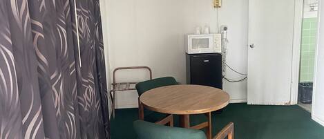 Habitación pequeña con una mesa redonda de madera y dos sillas, un microondas, una lámpara pequeña y una puerta que da al baño.