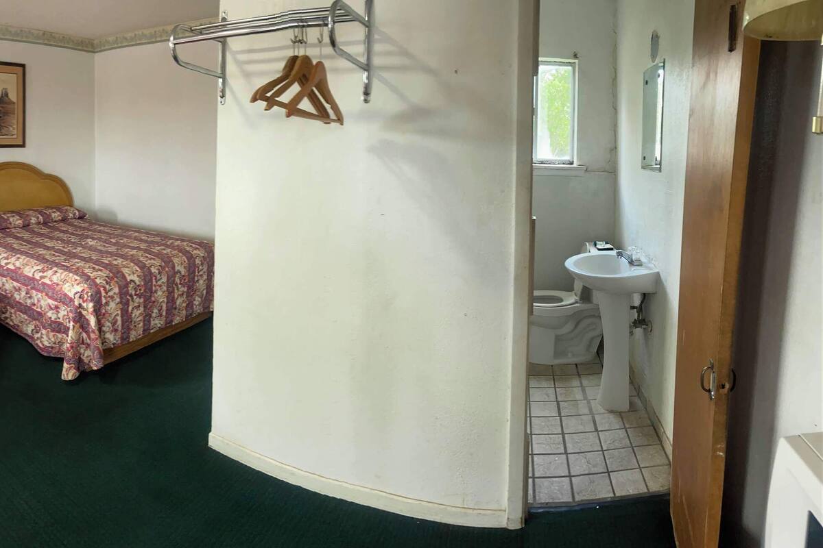 A room with three beds, a small table, a chair, a lamp, and a bathroom with a sink and toilet.