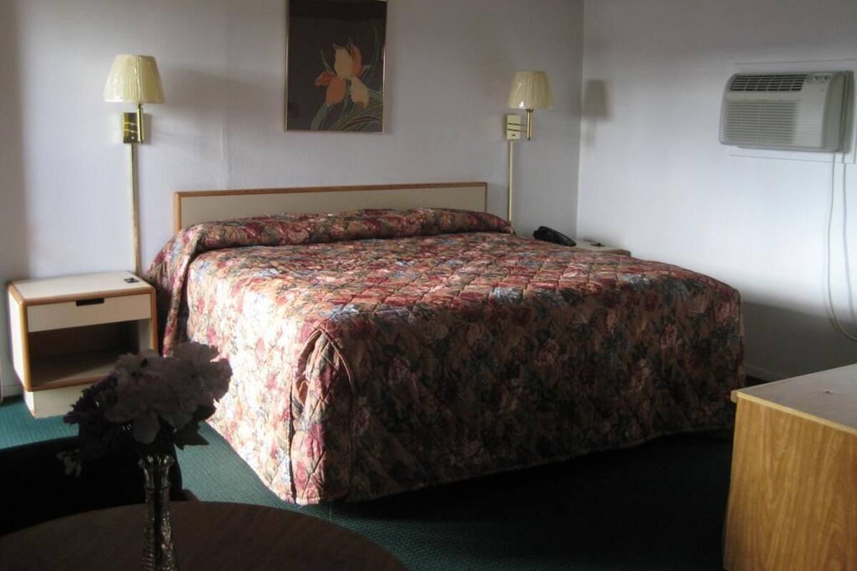 A bedroom with a bed, two bedside tables, a nightstand, a desk, a lamp, a vase with flowers, a painting, and an air conditioning unit.