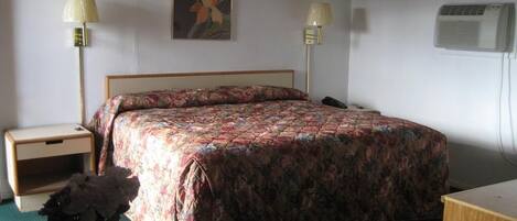 A bedroom with a bed, two bedside tables, a nightstand, a desk, a lamp, a vase with flowers, a painting, and an air conditioning unit.