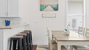 Una cocina con armarios blancos, encimera de granito y una cafetera. Hay una zona de comedor con una mesa y sillas de madera, y un cuadro de un muelle en la pared.