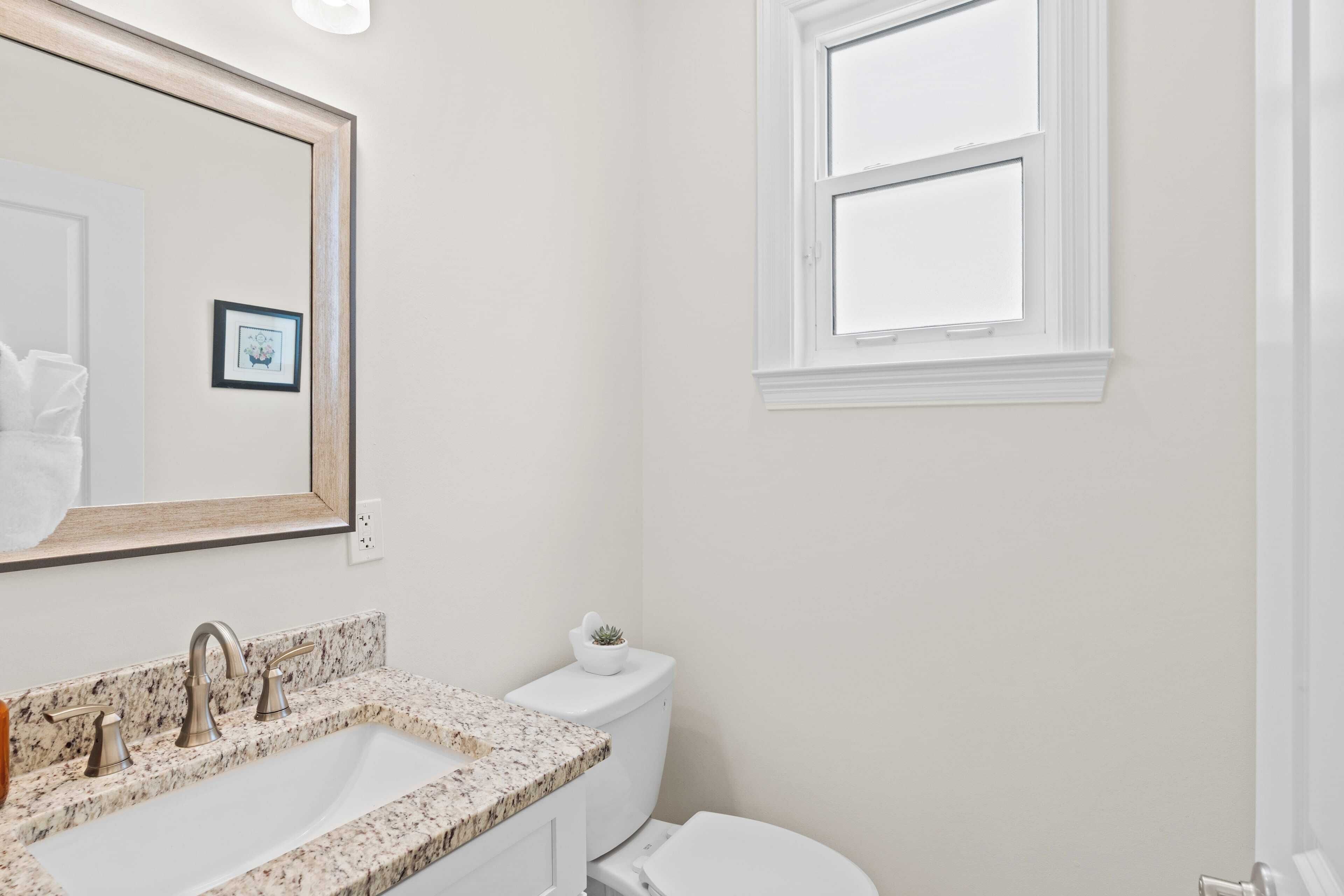 A bathroom with a sink, mirror, toilet, and window.
