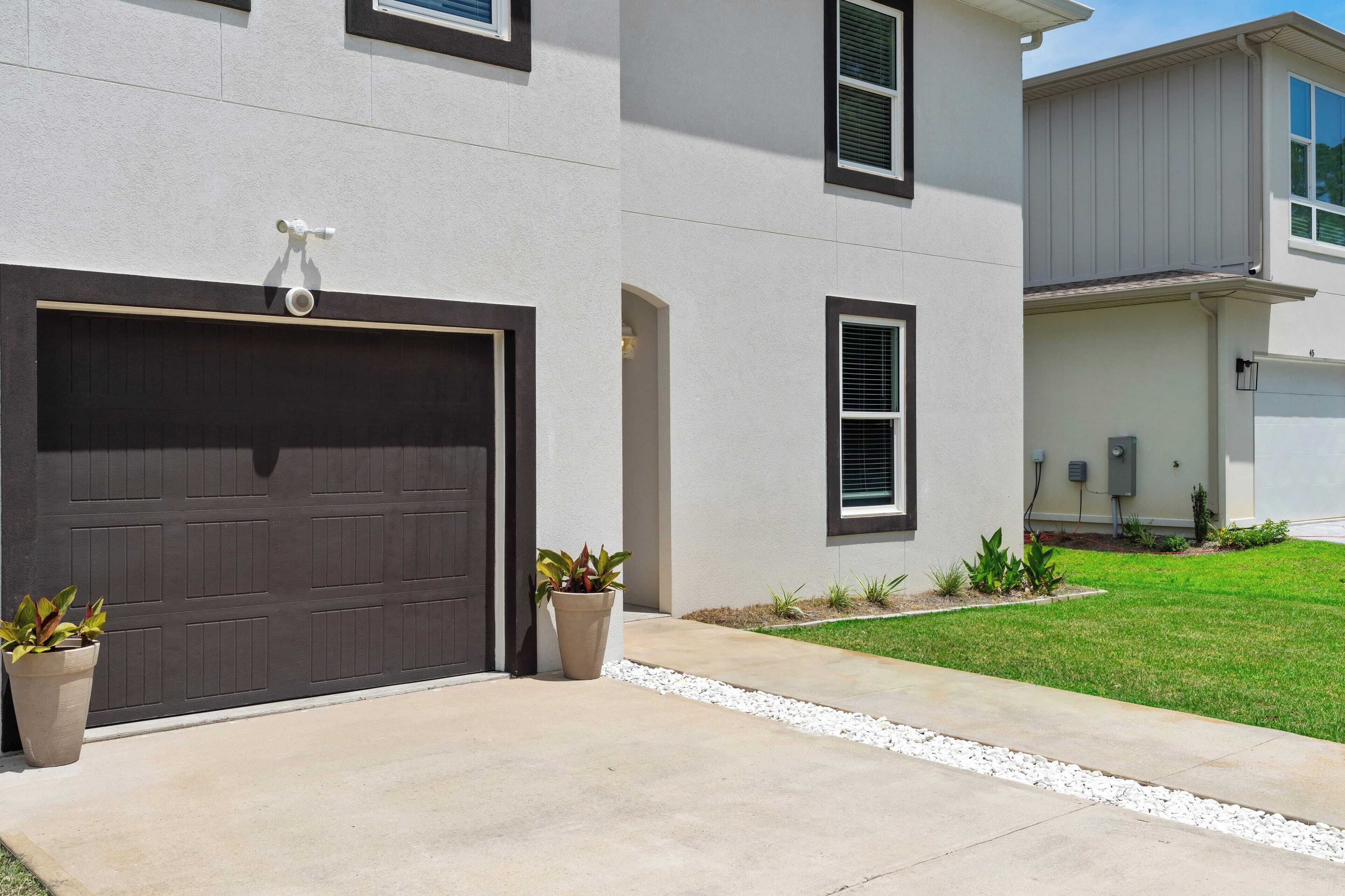 A modern two-story house with a dark garage door, a small front yard with a gravel pathway, and two potted plants.
