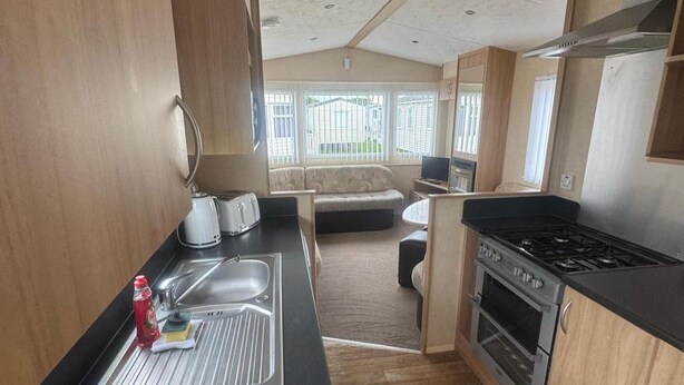 A compact kitchenette with a sink, toaster, and stove. A sofa and TV are visible in the background. - 3 Bed Caravan - Sleeps 8 - Pets - Parking (Rhyl)