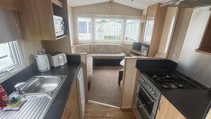 A compact kitchen with a sink, toaster, and stove, a small dining area with a sofa, and large windows offering a view of the outside. - 3 Bed Caravan - Sleeps 8 - Pets - Parking (Rhyl)