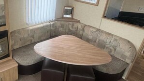 A compact RV interior with a curved sofa, a central table, and a small kitchenette area. - 3 Bed Caravan - Sleeps 8 - Pets - Parking (Rhyl)