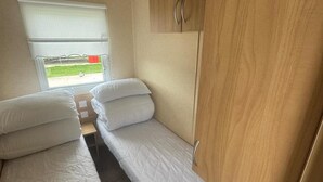 A compact RV bedroom with two beds, a window with a view, and a wooden cabinet. - 3 Bed Caravan - Sleeps 8 - Pets - Parking (Rhyl)