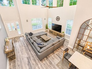 A spacious living room with a sectional sofa, a flat-screen TV, and a dining area with a table and chairs. - The Woodlands Gem | Heated Pool+Spa | EV Charging | Baby Crib | Ping Pong | Grill (The Woodlands)