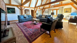 Ein geräumiges Wohnzimmer mit hohen Decken und freiliegenden Holzbalken, ausgestattet mit Sofas, Sesseln, einem Couchtisch und einem gemusterten Teppich.