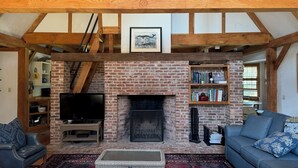 Ein rustikales Wohnzimmer mit freiliegenden Holzbalken, einem Kamin aus Ziegeln, einem Fernseher, einer Couch und einem Bücherregal.