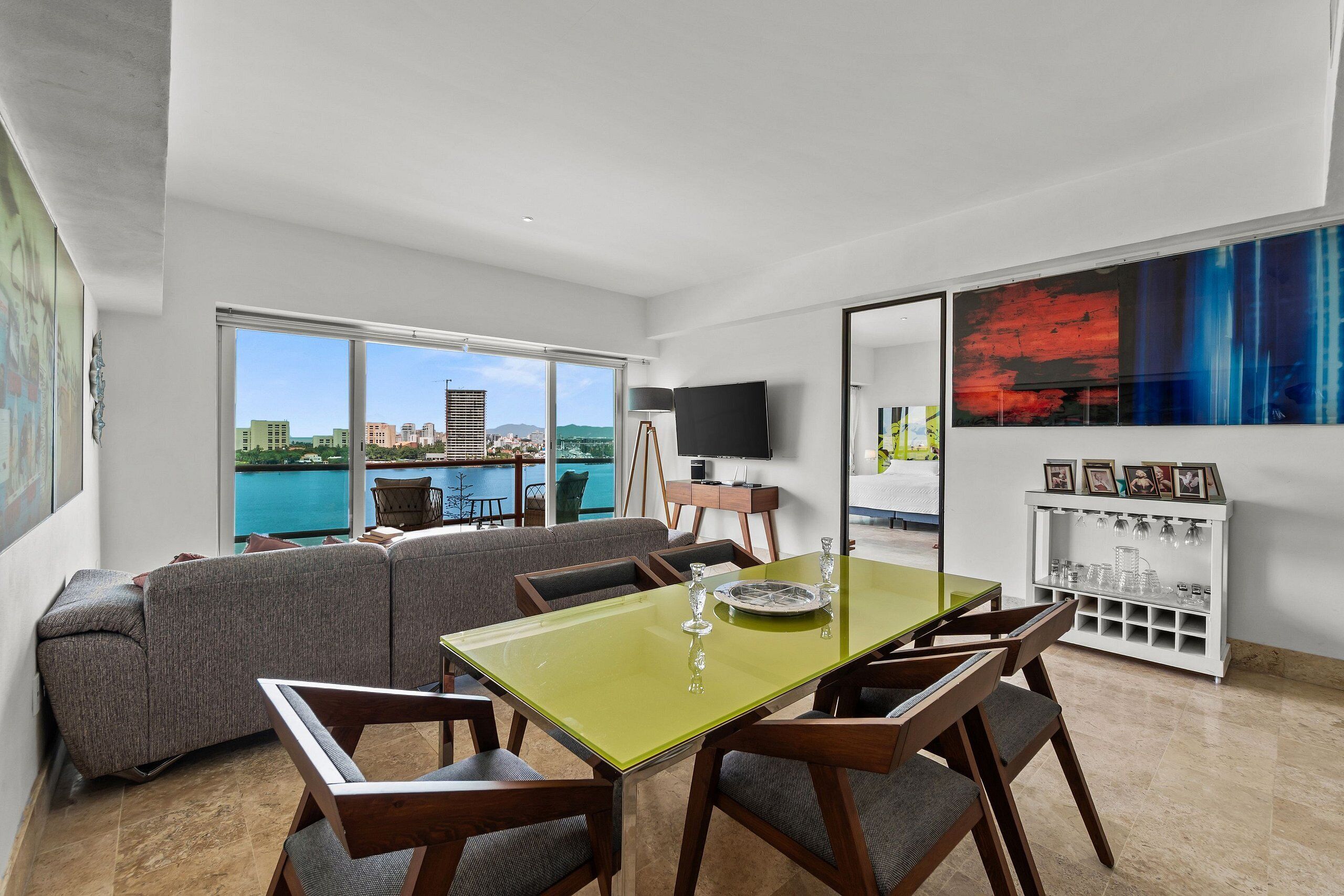 A modern living room with a dining area, large windows, a flat-screen TV, and a view of the city.