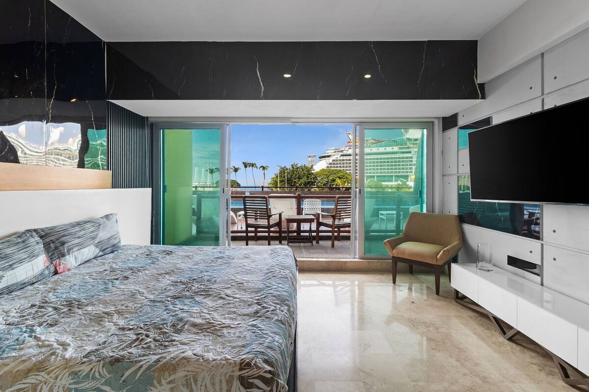 A modern hotel room with a bed, a TV, a balcony with a view of a cruise ship, and a dining area.