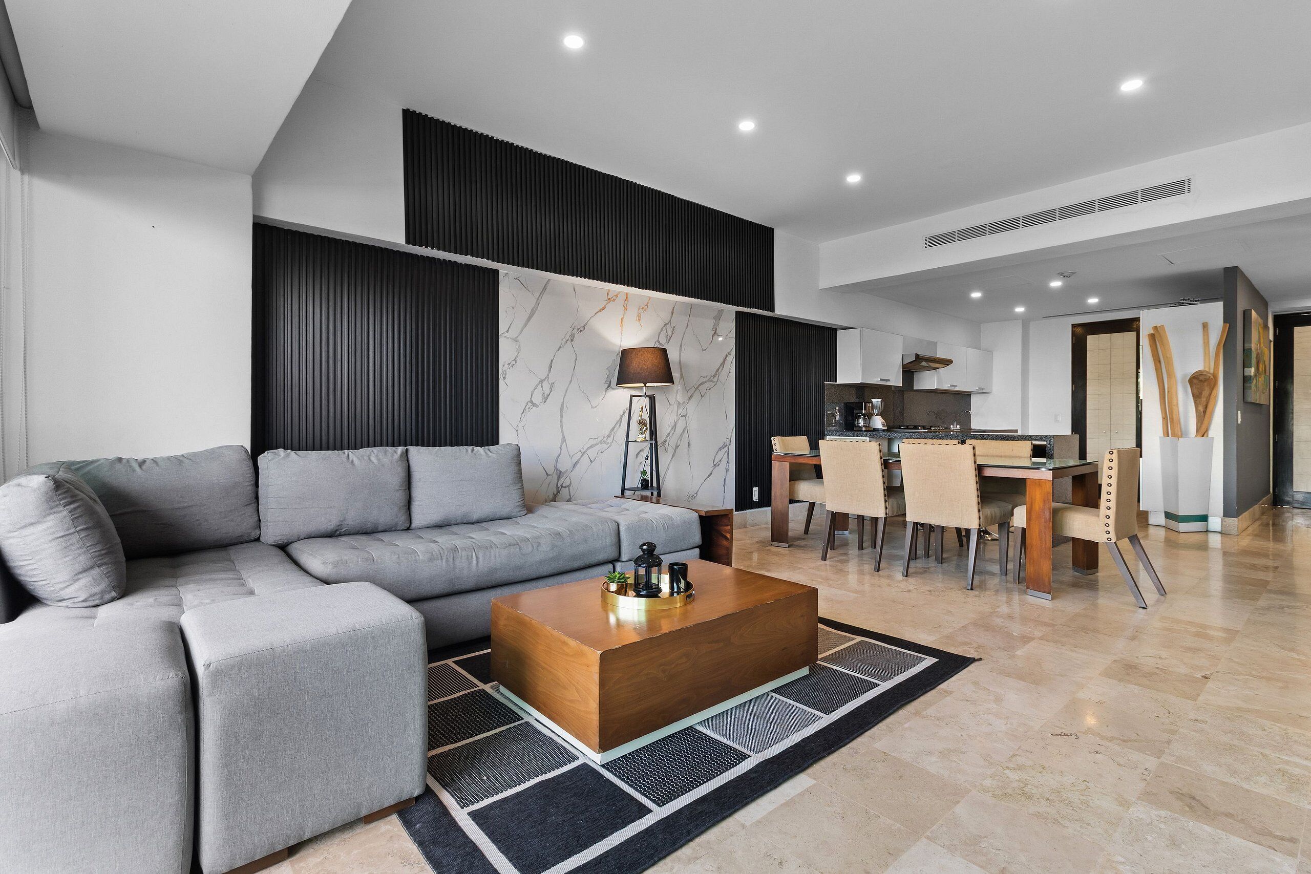 Una sala de estar moderna con un sofá seccional gris, una mesa de centro de madera y un comedor con una cocina al fondo.