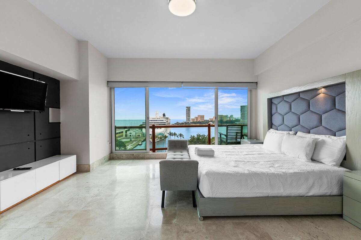 A modern bedroom with a large bed, a TV, and a balcony with a city view.