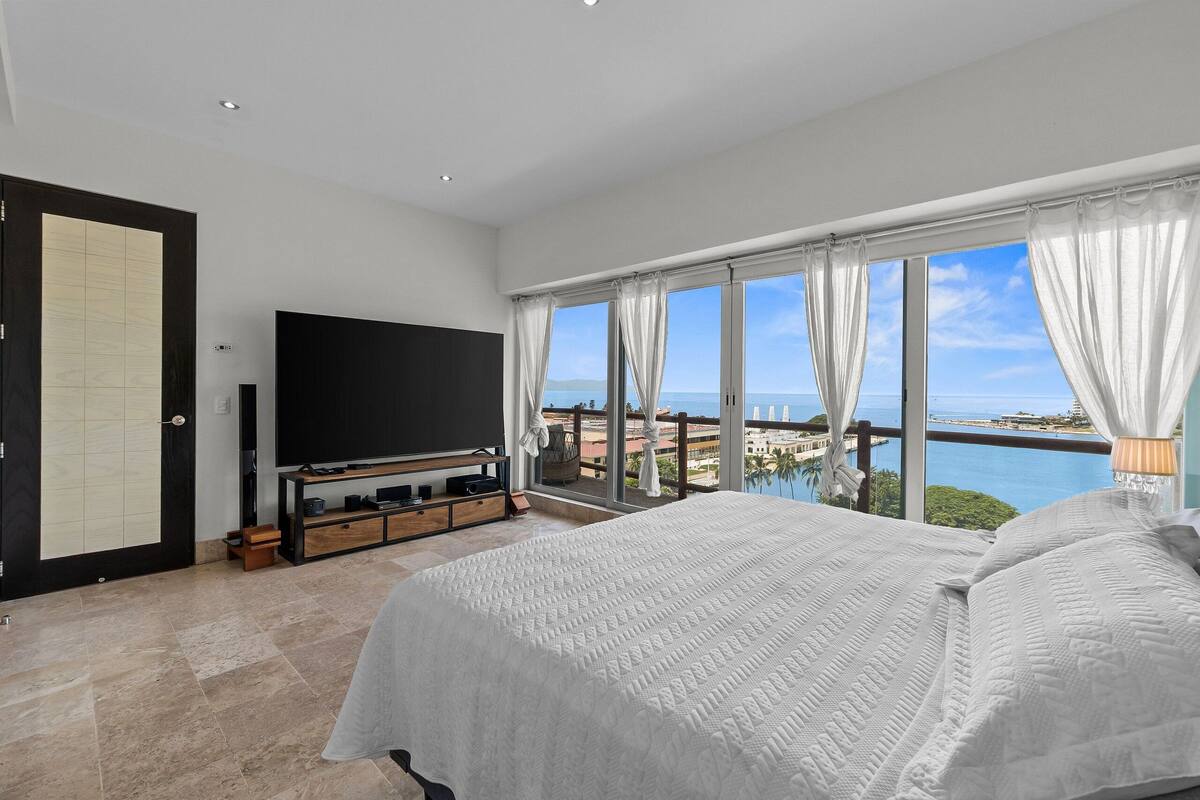 A bedroom with a large bed, a television, and a balcony with a view of the sea.