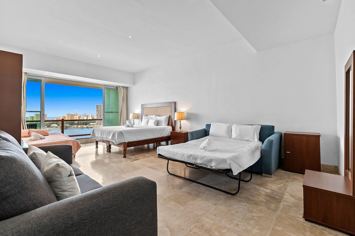 A hotel room with two beds, a sofa, a desk, and a view of the city.