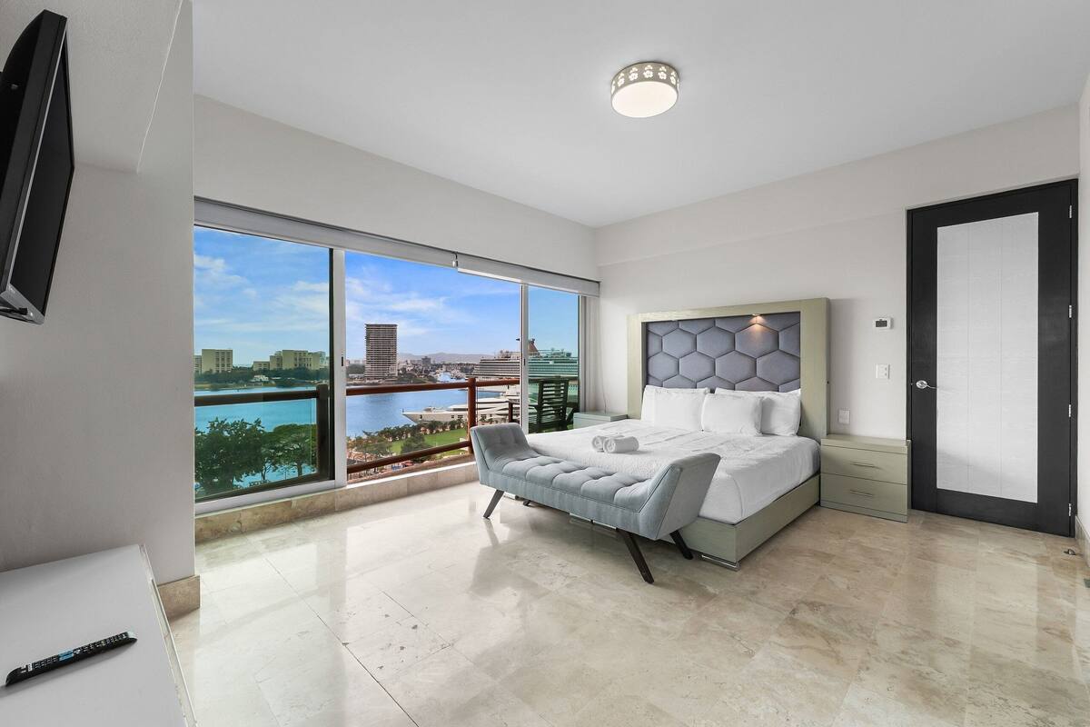 A modern bedroom with a large window, a bed, a bedside table, and a TV.