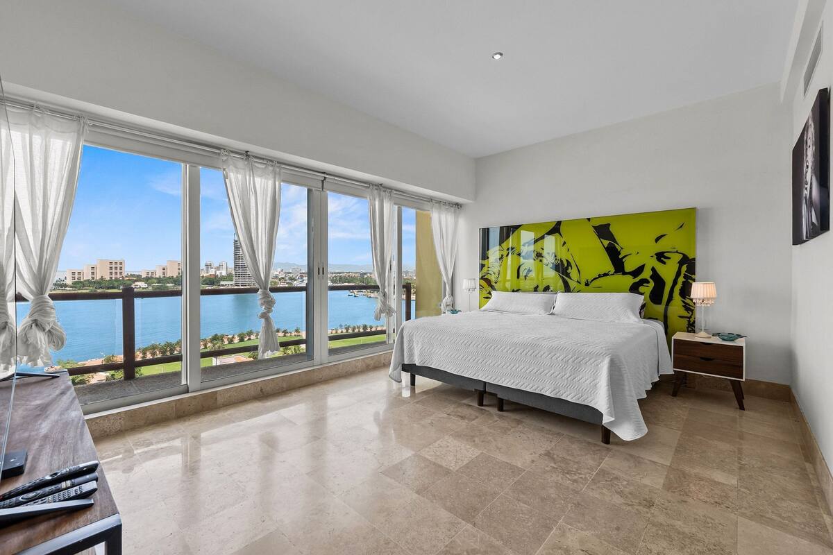 A bedroom with a large bed, a nightstand, a lamp, and a view of a body of water and buildings.