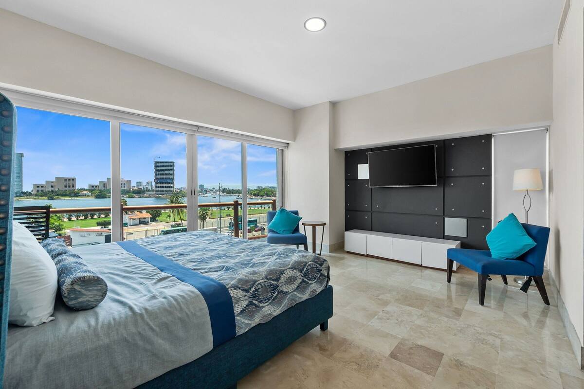 A modern hotel room with a large bed, a TV, a seating area, and a balcony with a city view.