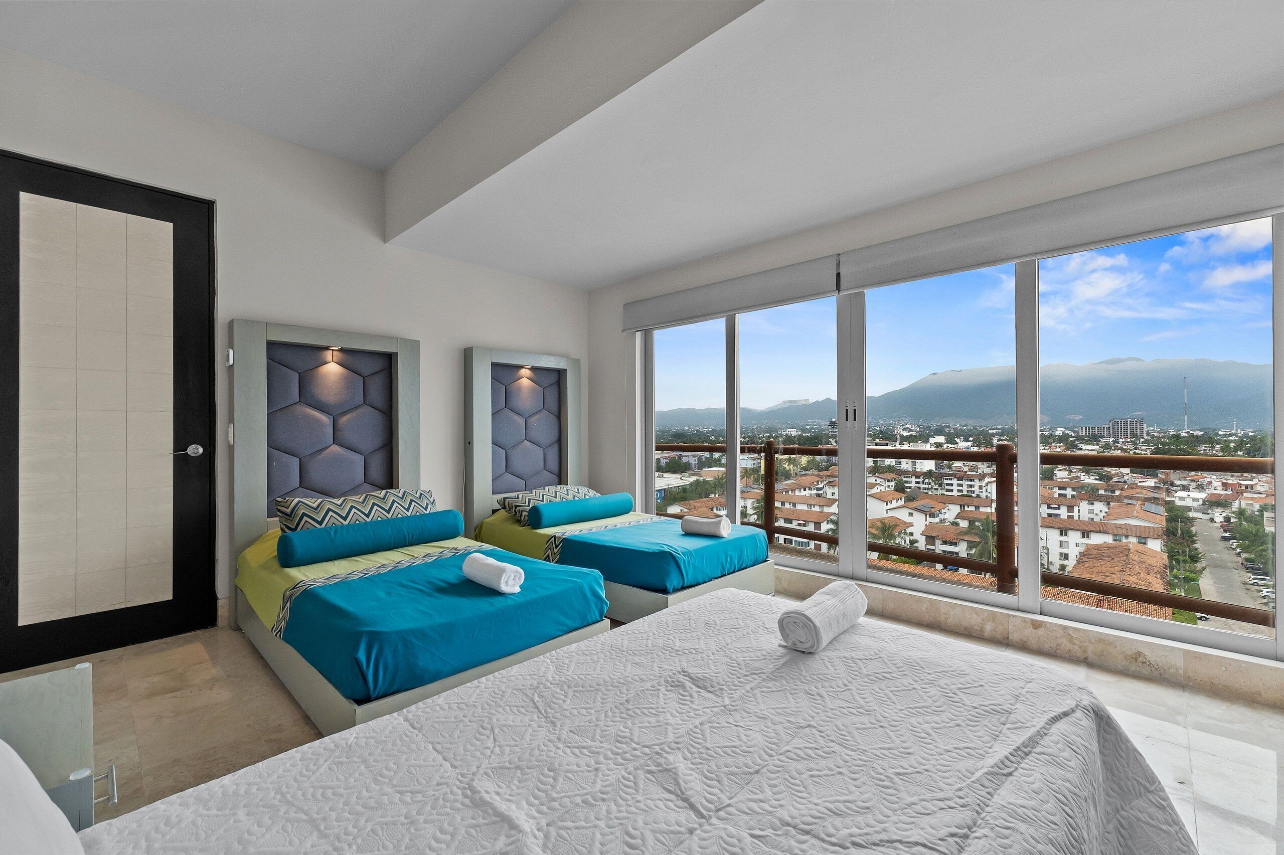 A hotel room with two beds, a large window with a city view, and a door.