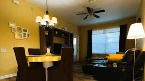 A living room with a sofa, a dining table with chairs, a ceiling fan, and a window with curtains.
