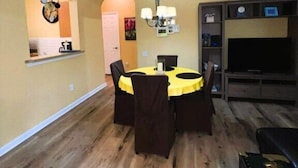 A dining area with a yellow table and chairs, a chandelier, and a TV stand with a flat-screen TV.