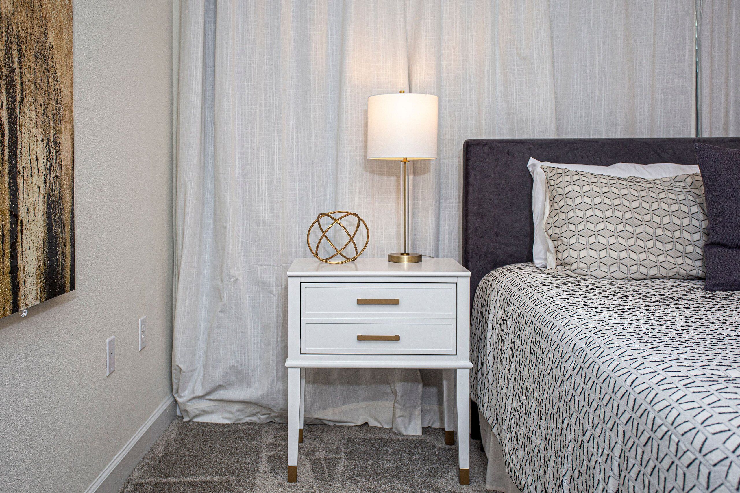 A neatly arranged bedroom with a white bedside table, a lamp, a patterned bedspread, and a dark headboard.
