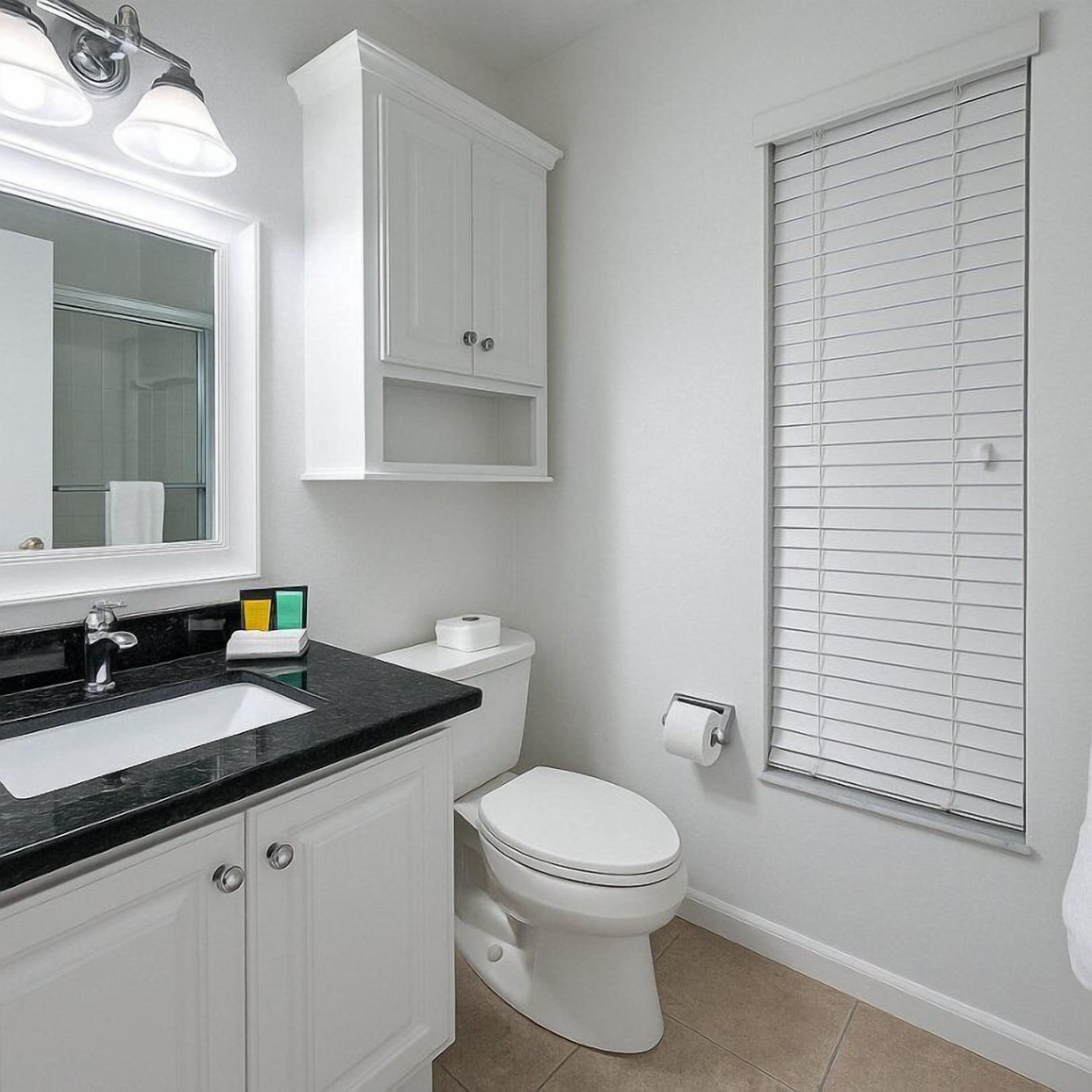 A bathroom with a white sink, mirror, and cabinet, a toilet, and a window with blinds.