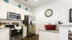 Een moderne keuken met witte kasten, roestvrijstalen apparatuur, een magnetron, oven en koelkast. Een klok hangt aan de muur en een rode broodrooster staat op het aanrecht.