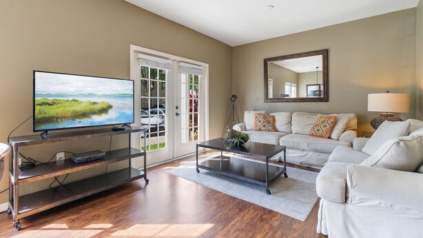 A living room with a flat-screen TV on a metal stand, a white sofa, a coffee table, and a large mirror on the wall.