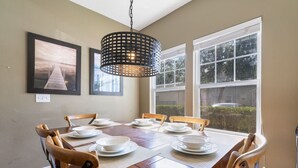 A dining room with a wooden table set for six, surrounded by chairs, with a large window and a hanging light fixture.