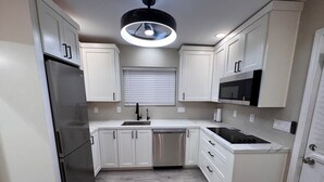 Una cocina compacta con armarios blancos, un refrigerador de acero inoxidable, lavavajillas, microondas y una lámpara sobre la encimera.